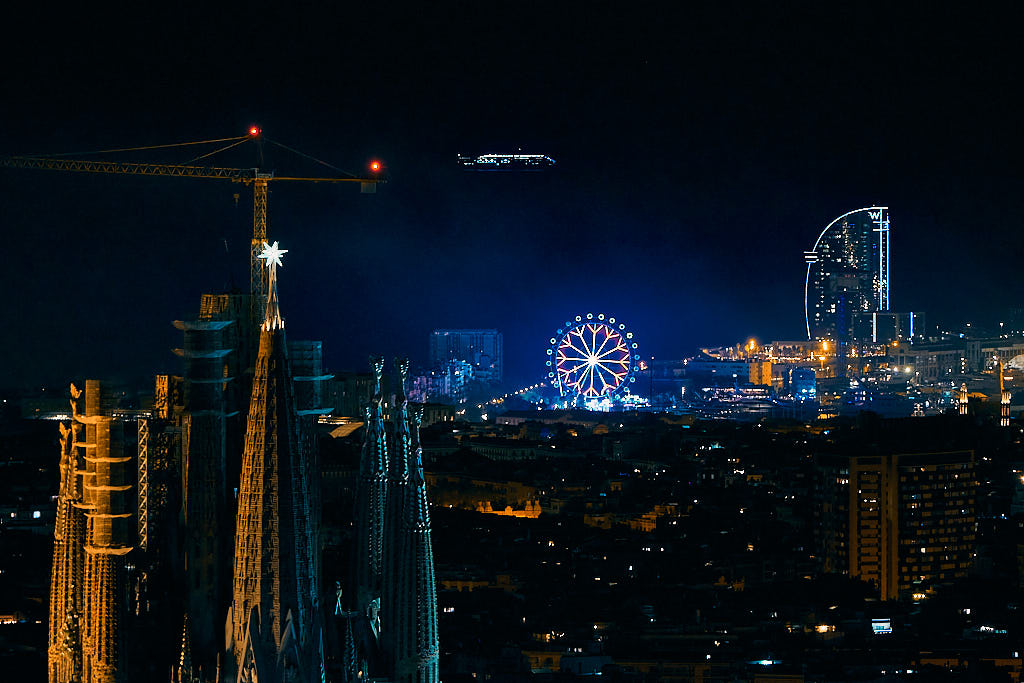 Un creuer que acaba de salpar del Port de Barcelona sembla suspès en l'aire per l'efecte òptic de la nit. En primer terme, la Sagrada Família. Al fons, l'Hotel W i la nòria de les Festes de la Mercè 2022.