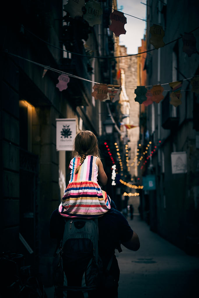 Una nena amb un vestit de molts colors es passeja sobre les espatlles del pare pel carrer dels Banys Vells, en el barri del Born de Barcelona.