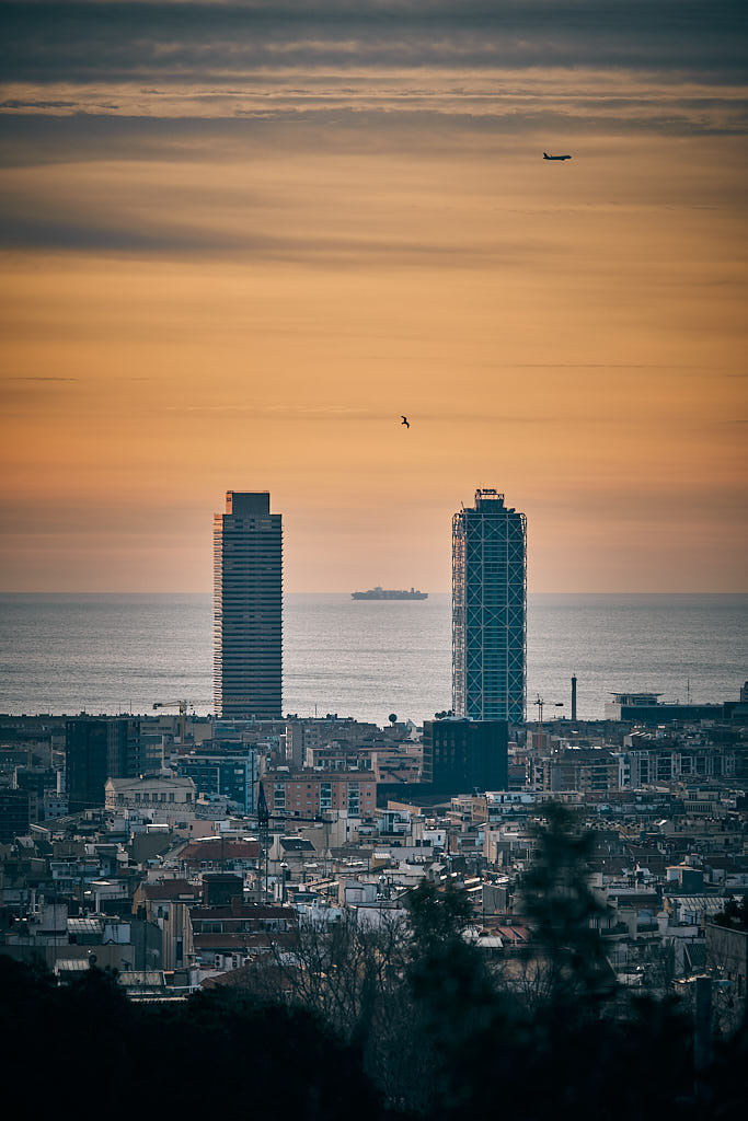 Un avió, un vaixell i una gavina es mouen rere les Torres Bessones de la Vila Olímpica del Poblenou de Barcelona. A l'esquerra, la Torre Mapfre; a la dreta, l'Hotel Arts.